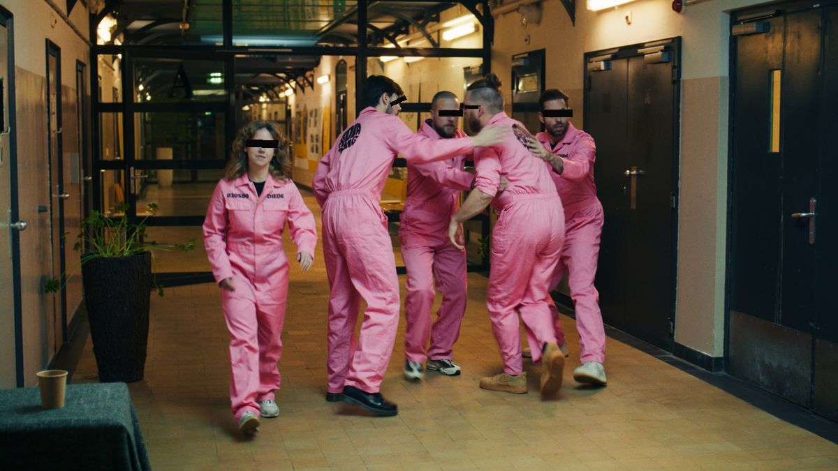 Mensen in roze overalls in een gevangenisgang, met zwarte balkjes over de ogen. Still uit een MediaTuig-commercial, 2024.