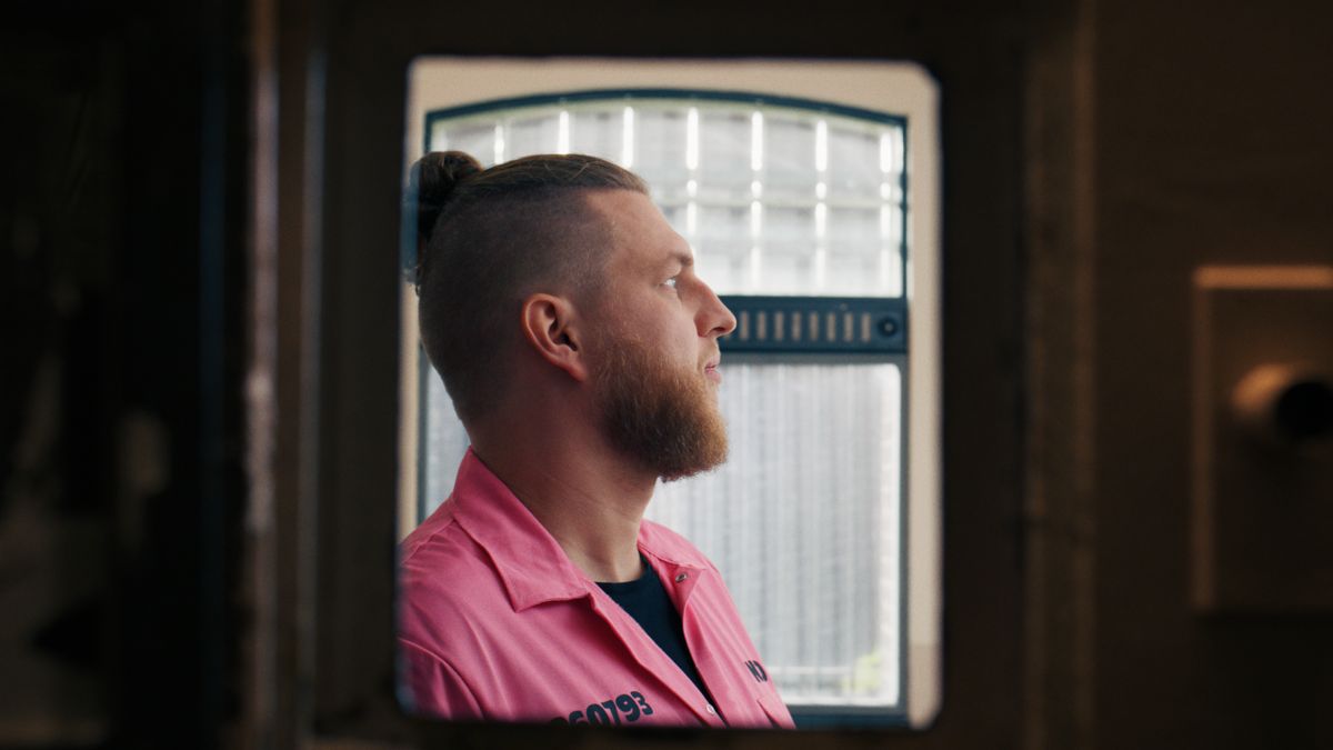 Een man in een roze overall kijkt door een celraam. Still uit een MediaTuig-commercial, 2024.